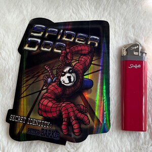 May include: A holographic sticker featuring a cartoon dog dressed as Spider-Man. The sticker has the text "Spider Dog" and "Secret Identity: Peter Barker".