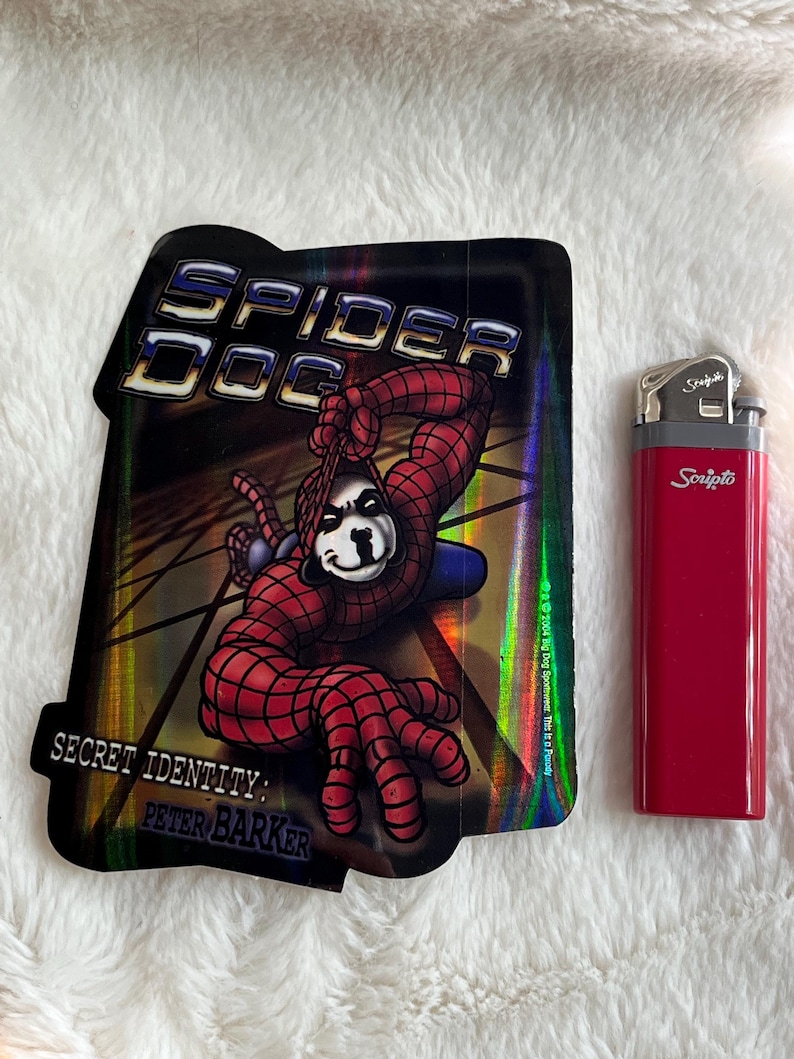 May include: A holographic sticker featuring a cartoon dog dressed as Spider-Man. The sticker has the text "Spider Dog" and "Secret Identity: Peter Barker".