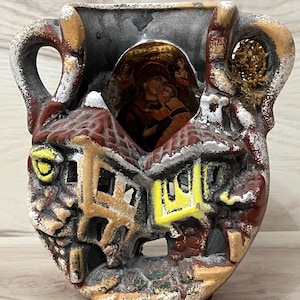 May include: A ceramic vase with a religious icon, featuring a depiction of Mary and Jesus. The vase has a unique design with a village scene, incorporating red, brown, and yellow hues. It has handles and decorative cutouts.