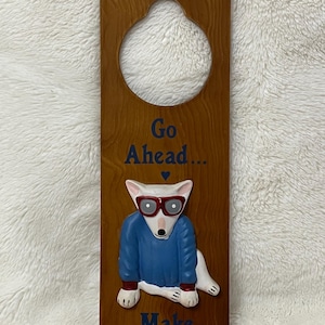 May include: Wooden door hanger with a cutout at the top. The brown hanger features a white dog wearing glasses and a blue sweater. Text reads "Go Ahead... Make My Bed!"