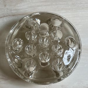 May include: Clear glass flower frog with multiple circular openings. The round frog has a shallow dish shape with a series of holes designed to hold flower stems in place for floral arrangements. The glass is transparent.