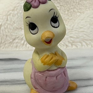 May include: A ceramic figurine of a cartoon chick with a flower crown and a pink apron. The chick has large, expressive eyes, a yellow beak, and is posed with its arms crossed. The figurine is painted in soft pastel colors.
