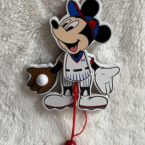 May include: A Mickey Mouse baseball-themed ornament. Mickey is wearing a baseball uniform, a blue and red cap, and holding a baseball glove with a white baseball. The ornament has a red string for hanging and a red bead.