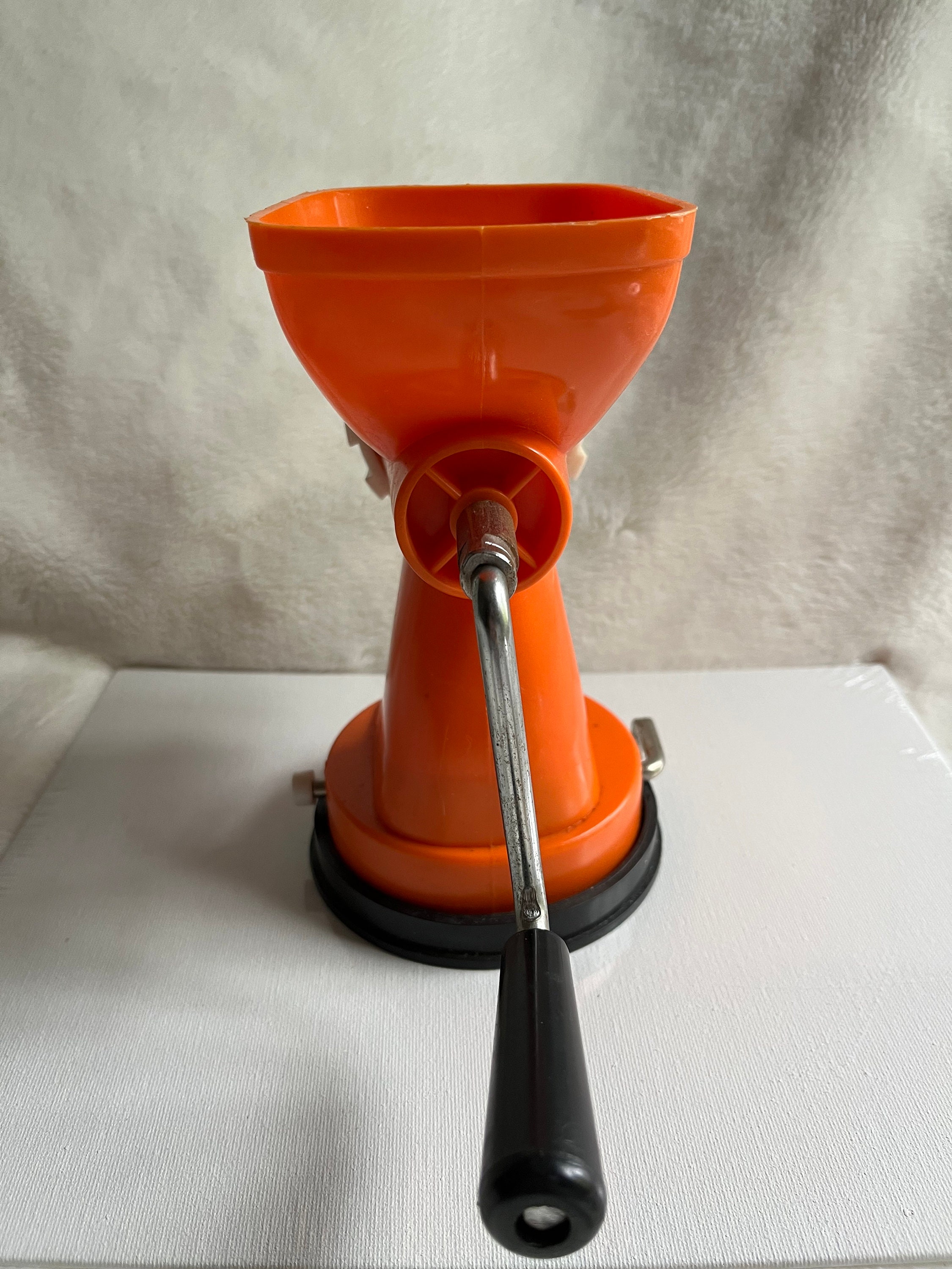 A bright orange meat grinder with a black base and a metal handle is placed on a white surface, against a neutral background.