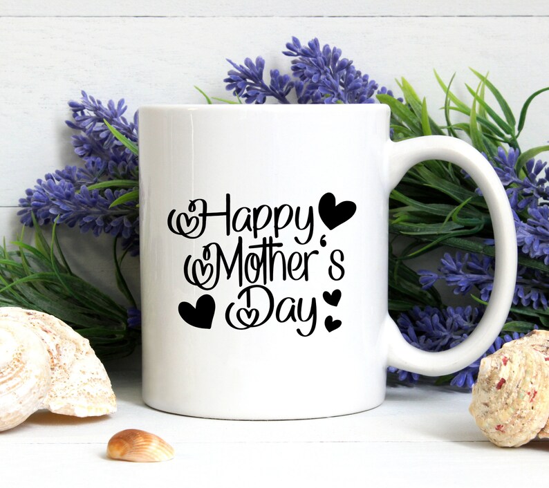 Puede incluir: Taza de cer&aacute;mica blanca con un dise&ntilde;o negro que dice "Feliz D&iacute;a de la Madre" con corazones alrededor del texto.