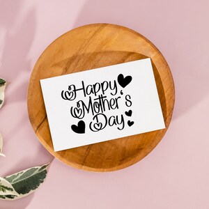 Puede incluir: Una tarjeta blanca con texto negro que dice "Happy Mother's Day" con peque&ntilde;os corazones que rodean el texto. La tarjeta est&aacute; sobre un plato de madera.