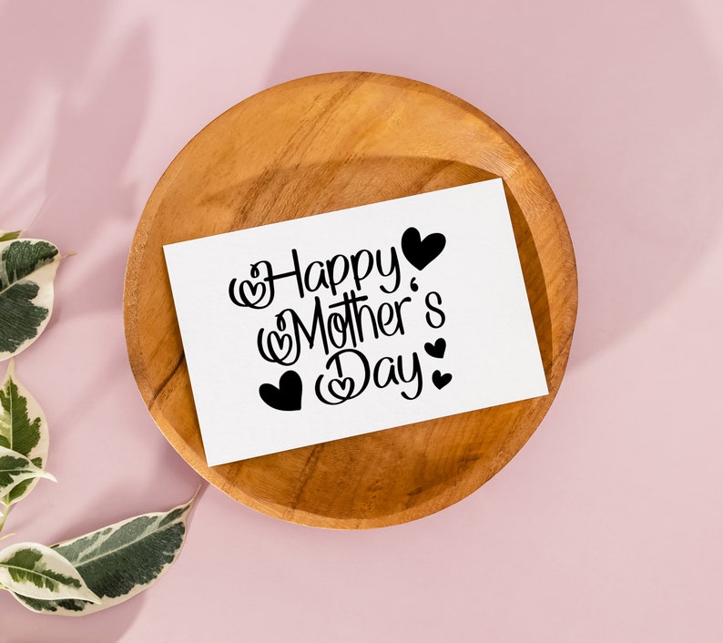 Puede incluir: Una tarjeta blanca con texto negro que dice "Happy Mother's Day" con peque&ntilde;os corazones que rodean el texto. La tarjeta est&aacute; sobre un plato de madera.