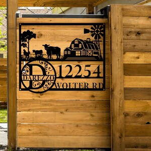 May include: Black metal address sign featuring a farm scene, including a barn, windmill, and cows, mounted on a wooden fence. The sign displays the address "12254 WOLTER RD" and the name "DAREZZO".