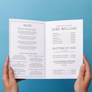 May include: An open booklet with the text "MUSIC" and "WHEN I AM BAPTIZED" on the left page, and "THE BAPTISM OF JAKE WILLIAM" on the right. The booklet is held open by two hands.