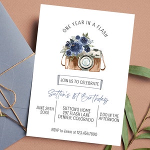 May include: A white invitation card with blue watercolor flowers and a vintage camera illustration. The text reads "One Year in a Flash" and "Sutton's 1st Birthday". The invitation includes the date, time, location, and RSVP information.