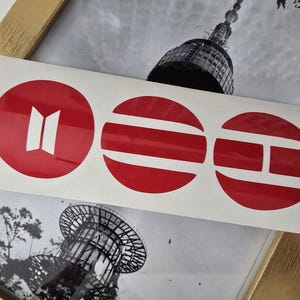 May include: A sheet of red and white stickers with three circular designs. The first circle has a white symbol, the second has horizontal stripes, and the third has a horizontal and vertical line design. The stickers are on a white background.