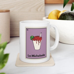 May include: White ceramic mug with a purple background and a cartoon illustration of a Michelada, a Mexican cocktail, with a lime wedge and a straw. The text "La Michelada" is printed below the illustration.