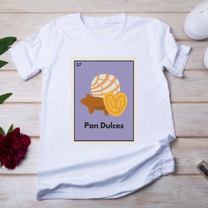May include: White t-shirt with a purple square graphic featuring a cartoon illustration of three Mexican pastries, a concha, a pan dulce, and a  'Pan Dulces' text label. The graphic also includes the number '27'.
