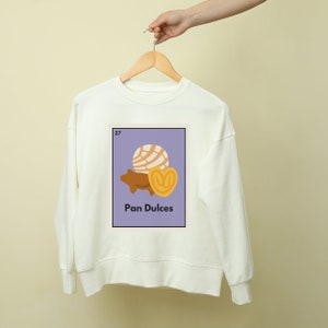 May include: White sweatshirt with a purple graphic of a Mexican sweet bread called "Pan Dulce" with the number 27 in the top left corner.