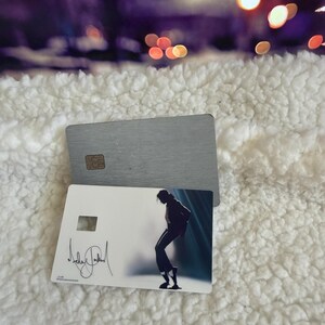 May include: Two rectangular cards, one with a brushed silver metallic finish and a gold chip, and another white card featuring a silhouette of a person dancing in a spotlight with a black signature printed on the left side of the card surface.