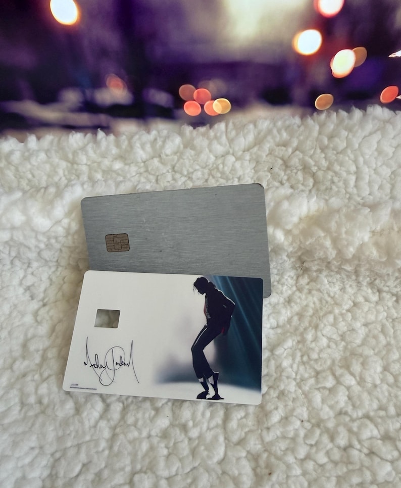 May include: Two rectangular cards, one with a brushed silver metallic finish and a gold chip, and another white card featuring a silhouette of a person dancing in a spotlight with a black signature printed on the left side of the card surface.