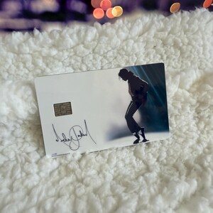 May include: A rectangular white credit card featuring a metallic chip on the left side. The card displays a black silhouette of a person dancing in a dynamic pose on the right, with a black signature printed in the lower left corner of the card.