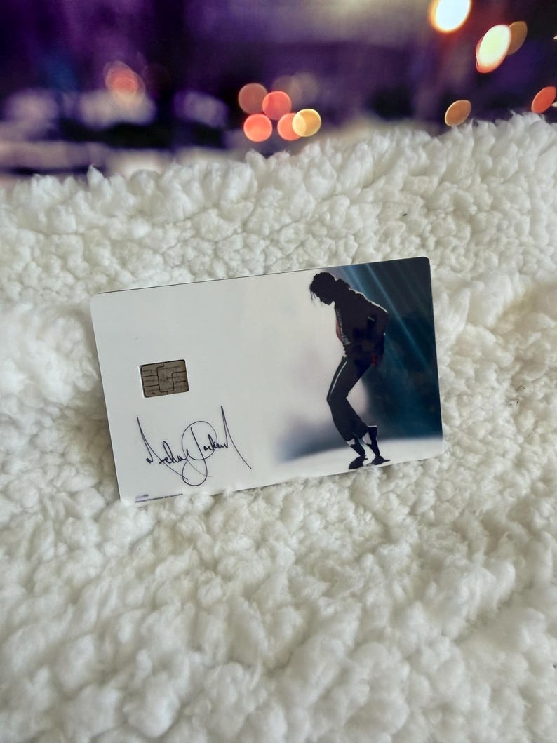 May include: A rectangular white credit card featuring a metallic chip on the left side. The card displays a black silhouette of a person dancing in a dynamic pose on the right, with a black signature printed in the lower left corner of the card.