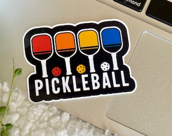 Pode incluir: Um adesivo preto e branco com quatro raquetes de pickleball coloridas e a palavra "PICKLEBALL" em letras brancas.