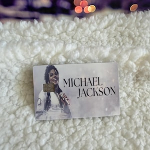May include: A rectangular credit card featuring a black and white photographic print of Michael Jackson holding a microphone. The card includes a gold-colored EMV chip on the left side and the text MICHAEL JACKSON printed in black on the right side.
