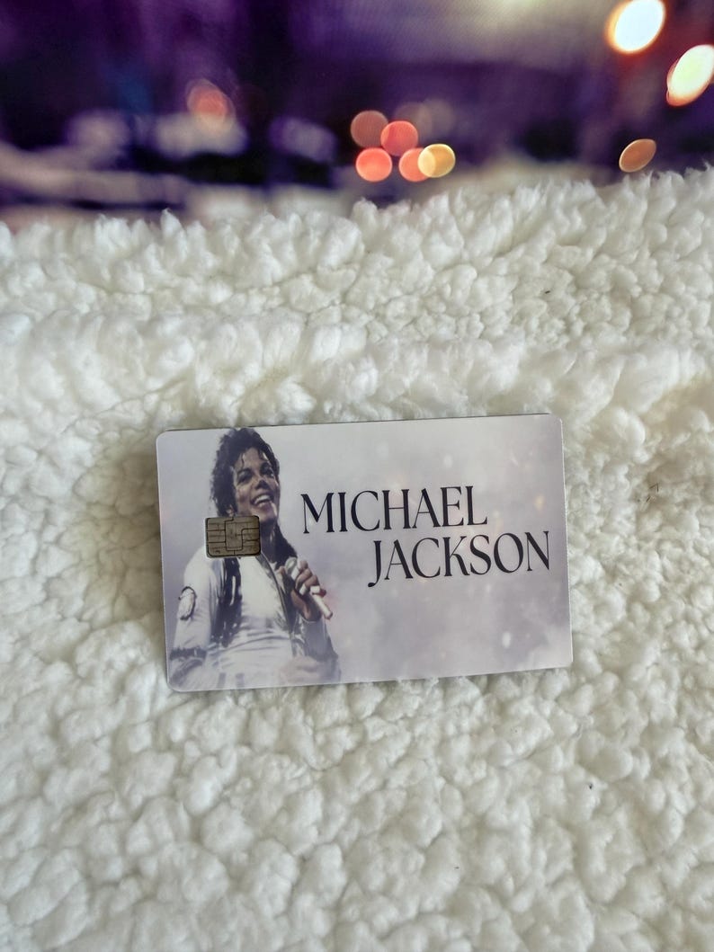 May include: A rectangular credit card featuring a black and white photographic print of Michael Jackson holding a microphone. The card includes a gold-colored EMV chip on the left side and the text MICHAEL JACKSON printed in black on the right side.