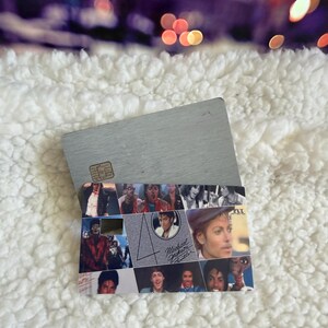 May include: A rectangular credit card featuring a collage of Michael Jackson images from the Thriller era. The design includes the number 40, the text Michael Jackson Thriller, and the P.Y.T. logo. The card has a metallic silver finish and a chip.