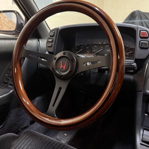 May include: A wooden steering wheel with a dark brown finish and black spokes. The center features a black emblem with a red "H" logo. The wheel is mounted in a car interior with a black dashboard and gray seat.