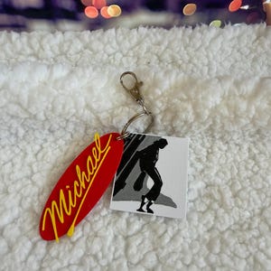 May include: A keychain featuring a red oval charm with the name Michael written in yellow script and a square white charm displaying a black and white silhouette illustration of a person dancing in a pose, attached to a silver metal lobster clasp.