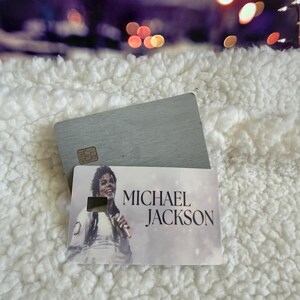 May include: A rectangular card featuring a printed image of Michael Jackson holding a microphone, with the text MICHAEL JACKSON printed in black. The card is layered over a plain, brushed silver-toned metal card with a visible gold-colored chip.