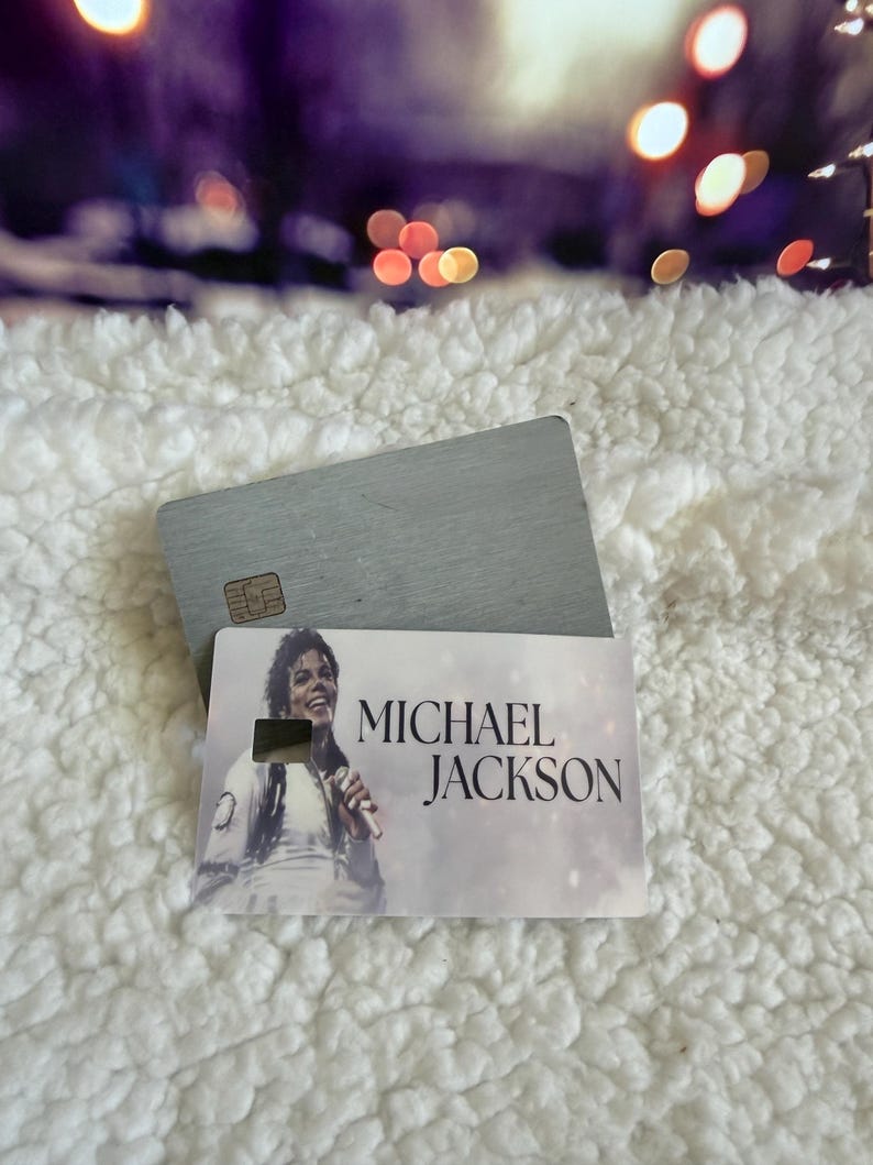 May include: A rectangular card featuring a printed image of Michael Jackson holding a microphone, with the text MICHAEL JACKSON printed in black. The card is layered over a plain, brushed silver-toned metal card with a visible gold-colored chip.