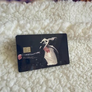 May include: A rectangular credit card featuring a dark background with a printed image of a performer wearing a black sequined jacket, a white shirt, and a black fedora hat. The card includes a gold-colored EMV chip on the left side.