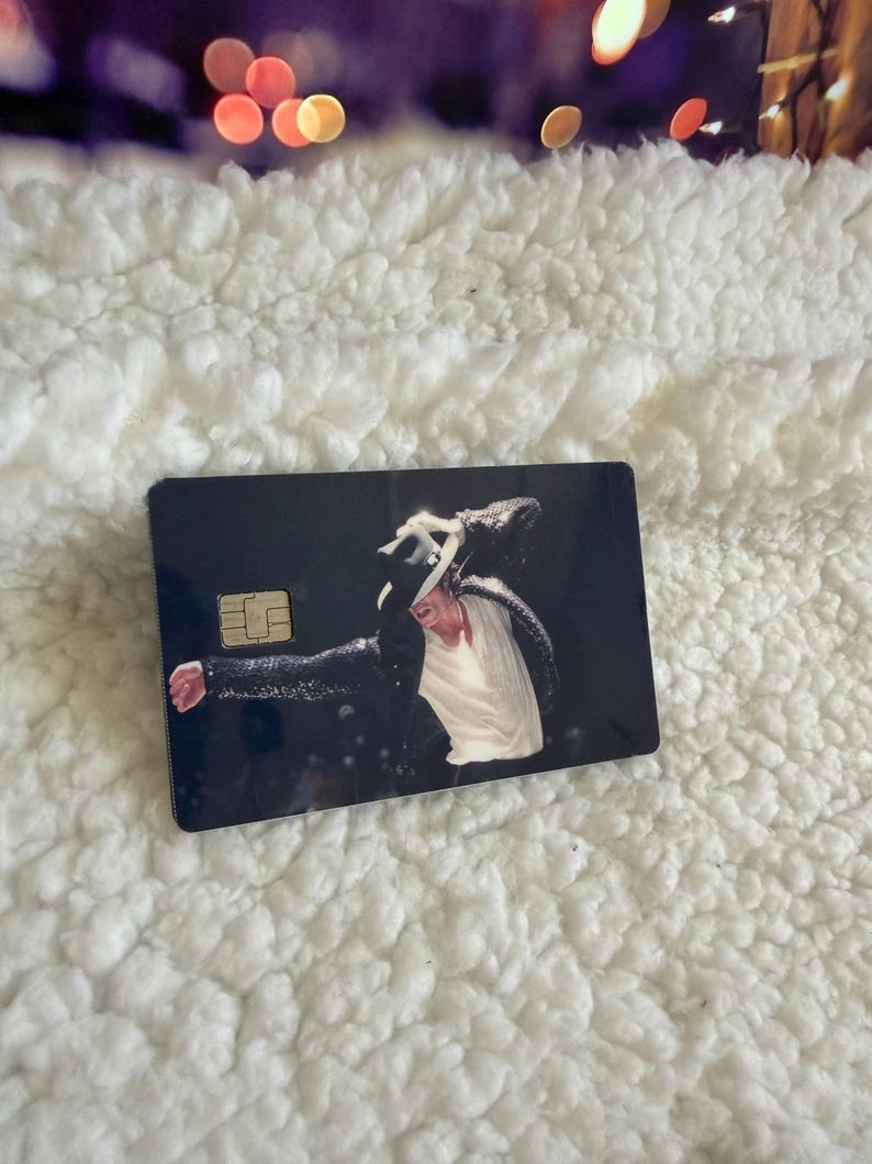 May include: A rectangular credit card featuring a dark background with a printed image of a performer wearing a black sequined jacket, a white shirt, and a black fedora hat. The card includes a gold-colored EMV chip on the left side.
