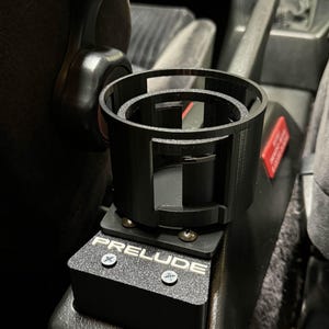 May include: Black car cup holder with the word "PRELUDE" printed on it. The cup holder is attached to the side of a car seat.