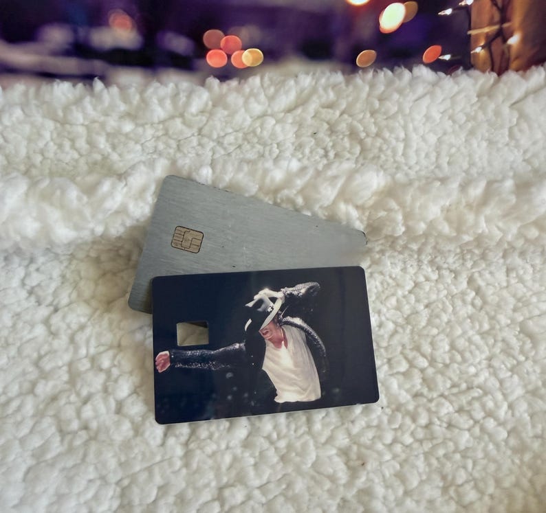 May include: A rectangular credit card featuring a printed image of a performer wearing a black sequined jacket, a white shirt, and a black fedora hat. The card has a small square cutout on the left side and a metallic chip embedded on the top surface.