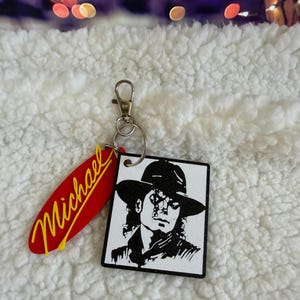 May include: A keychain featuring a square white pendant with a black graphic illustration of a person wearing a fedora hat, attached to a metal ring and clasp. A secondary red, surfboard-shaped pendant with the name Michael in yellow script is attached.