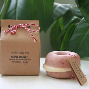 May include: A small, pink and white handmade soap shaped like a bagel. The soap is packaged in a brown paper bag with a red and white string tied around it. The bag has the text "Studio Happy Tree MINI BAGEL Raw Goat Milk with Madder Root Handmade Soap" printed on it.