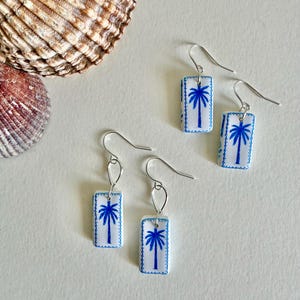 May include: Two pairs of rectangular drop earrings featuring a white base with a blue palm tree illustration and a decorative blue dotted border. Each earring hangs from a silver-toned metal hook, showcasing a clean, symmetrical design.