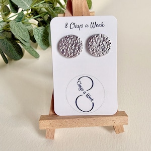 May include: Silver-coloured, textured stud earrings on a white card with the text "8 Clays a Week" and a logo. The card is displayed on a small wooden easel, with greenery in the background. The earrings are circular.