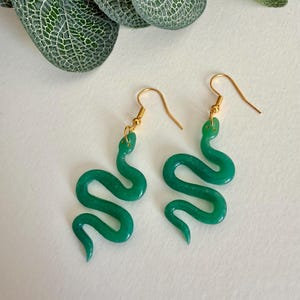 May include: A pair of drop earrings featuring translucent green snake-shaped charms suspended from gold-toned metal fishhook wires. The snakes are depicted in a curved, S-shaped posture with visible head details and a smooth, glossy finish.