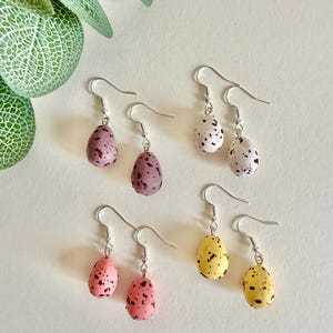 May include: Six pairs of egg-shaped earrings in shades of purple, white, pink, and yellow. Each earring features a speckled pattern and a silver hook. The earrings are presented on a white surface.