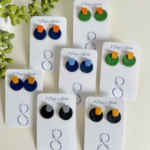 May include: A selection of handmade stud earrings in diverse colour combinations, such as navy blue with orange, green with orange, and black with grey. Each pair is presented on a white card with the text "8 Clays a Week".