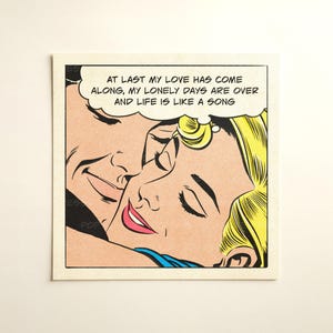 May include: A square print featuring a comic book style illustration of two people embracing with eyes closed. A speech bubble above them contains the text: AT LAST MY LOVE HAS COME ALONG, MY LONELY DAYS ARE OVER AND LIFE IS LIKE A SONG.