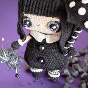 May include: A black and white crocheted doll with a black coat, white collar, and black and white striped leggings. The doll is holding a gray crocheted doll with a stitched X on its chest. The doll is standing on a purple surface with black rocks.