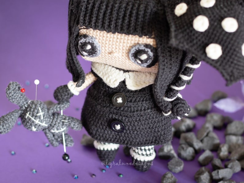 May include: A black and white crocheted doll with a black coat, white collar, and black and white striped leggings. The doll is holding a gray crocheted doll with a stitched X on its chest. The doll is standing on a purple surface with black rocks.