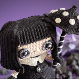 May include: A crocheted doll with black hair, a black dress, and a white collar. The doll is holding a black and white crocheted umbrella with white polka dots.