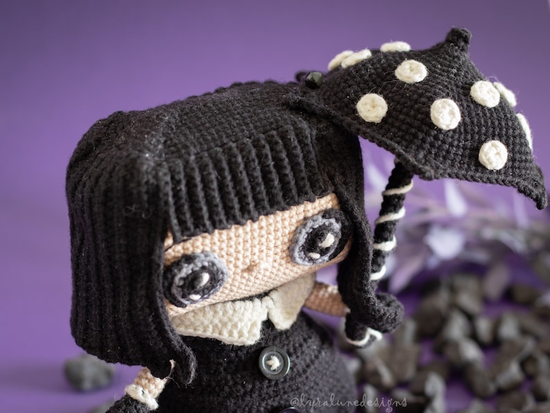 May include: A crocheted doll with black hair, a black dress, and a white collar. The doll is holding a black and white crocheted umbrella with white polka dots.