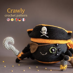 以下が含まれることがあります： 黒とオレンジの海賊帽をかぶった、かぎ針編みで作られたクモ。眼帯とドクロと交差した骨のデザインが施されています。クモは片手にフック、もう片手にランタンを持っています。クモは、周りに金色のキラキラが散らばった茶色の表面に座っています。