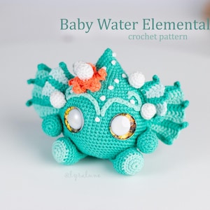 Könnte beinhalten: Eine türkisfarbene Häkelfigur mit großen weißen Augen, orangefarbenen und weißen Akzenten sowie flossenartigen seitlichen Anhängseln. Der Text Baby Water Elemental crochet pattern ist über dem Objekt und der Name @lyralune darunter.