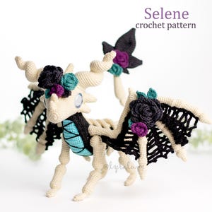 Op de afbeelding: Een gehaakte draak met een wit lichaam, zwarte vleugels en turquoise accenten. De draak heeft zwarte en paarse bloemen op zijn kop en vleugels. De draak staat op een wit oppervlak. "Selene crochet pattern" staat in de rechterbovenhoek van de afbeelding.