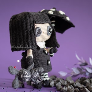May include: A black crocheted doll with long black hair, wearing a black dress and a white collar. The doll is holding a small black crocheted doll with an X on its chest. The doll is standing on a purple surface with black rocks and glitter.